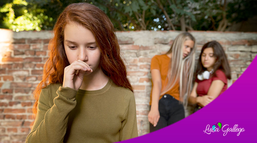 Una niña con expresión de preocupación y la mano cerca de la boca, sentada sola en un patio de ladrillos, mientras otras dos niñas conversan de espaldas a ella. La imagen representa la sensación de aislamiento y la ansiedad social en la infancia.
