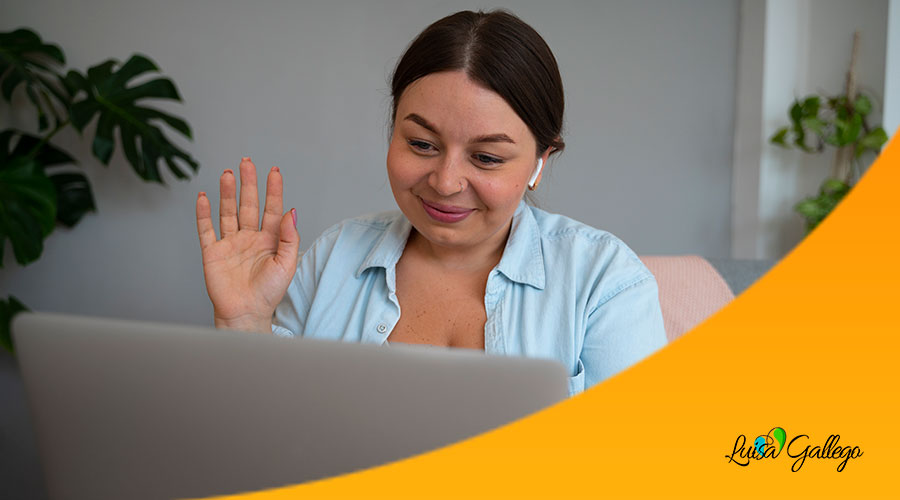 Una mujer sonriente saluda con la mano a la cámara de su portátil, con auriculares inalámbricos. La imagen refleja la comodidad y la accesibilidad de la terapia online desde casa.